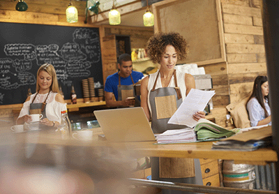a coffee shop owner is working on her website on the counter of her busy coffee shop. In the background two staff attend to the orders.