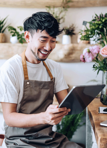 Asian male florist, owner of small business flower shop, using digital tablet while working on laptop against flowers and plants.