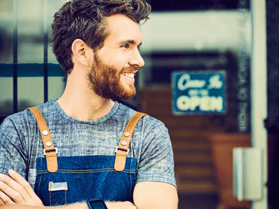 Smiling small business owner in apron looking away. Confident barista is outside cafe. He is standing arms crossed against window.